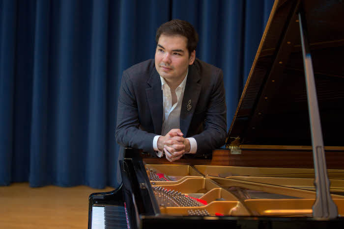 Alim leaning on the side of the piano towards the camera but looking off to his right. Photo credit