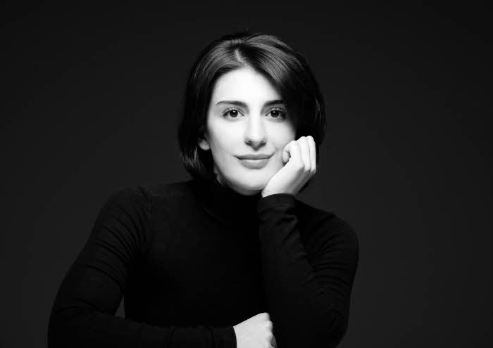Mariam shown with her right arm in front of her on - presumably - the top of a piano; the other arm is folded up and her chin rests on her left hand.